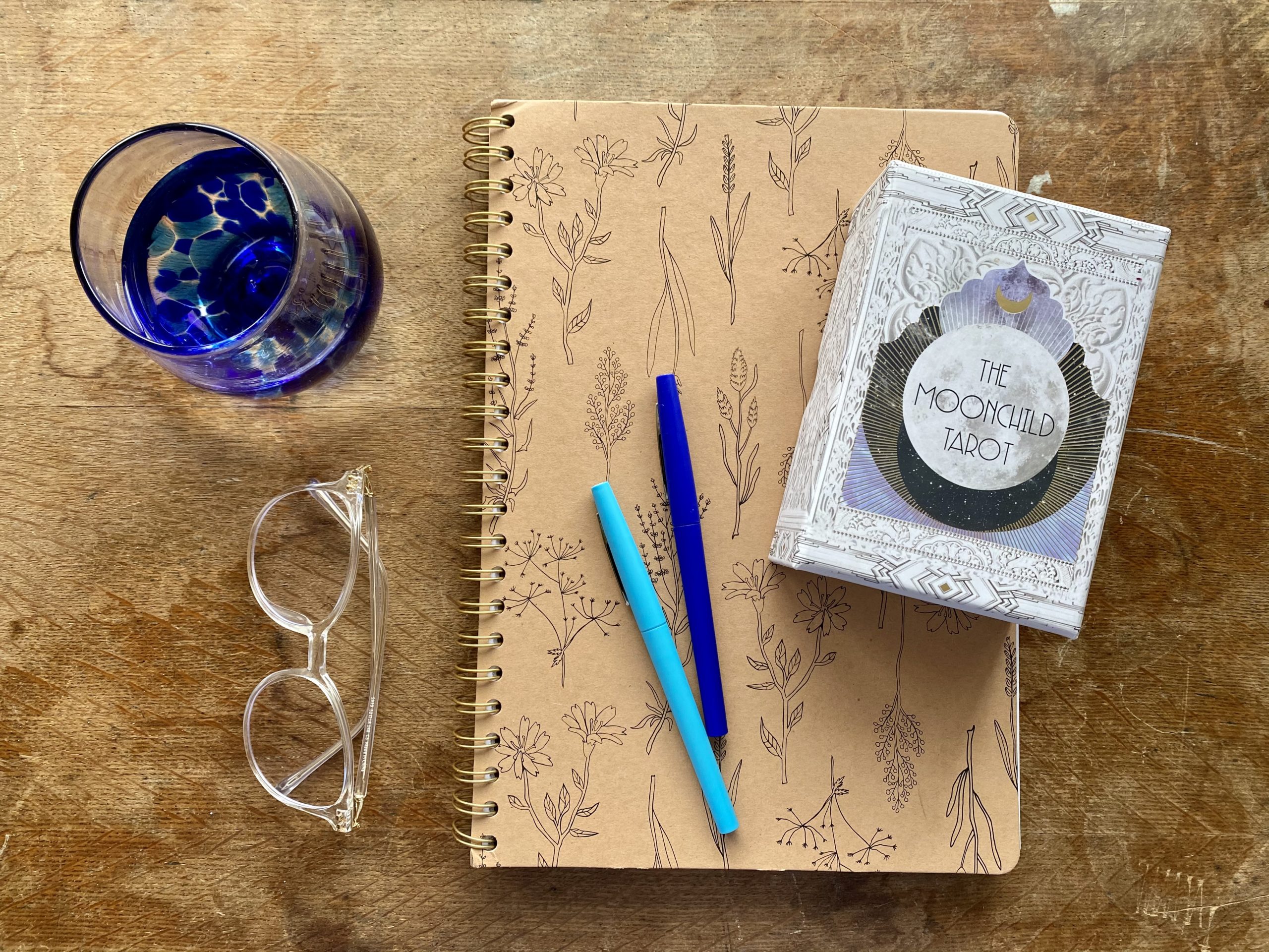 A journal rests on a desk with two blue markers, a pair of clear glasses, a blue water glass and a deck of tarot cards