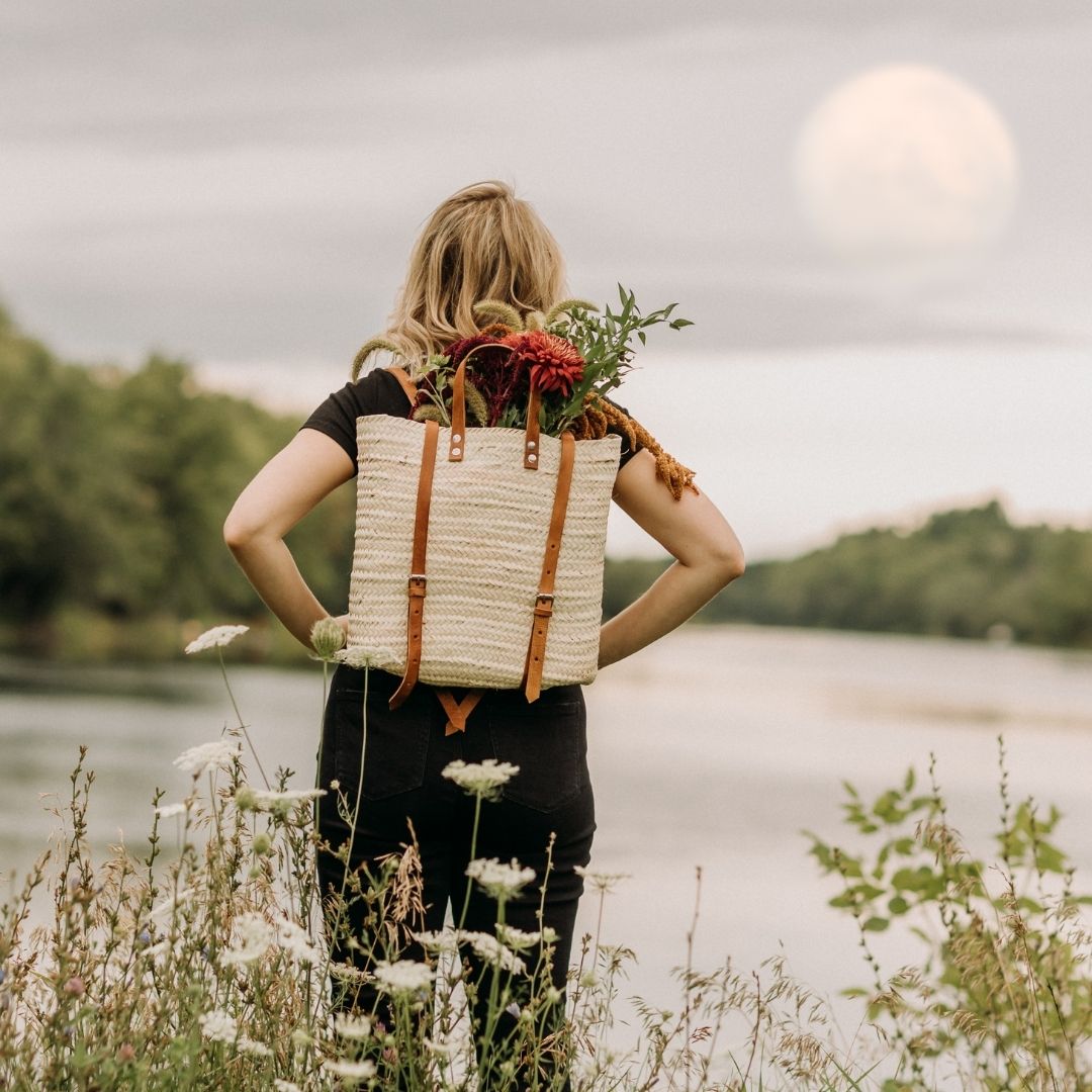 Lauren has a wicker backpack on with a bunch of flowers sticking out the top. She's overlooking a lake with the moon in the distance.