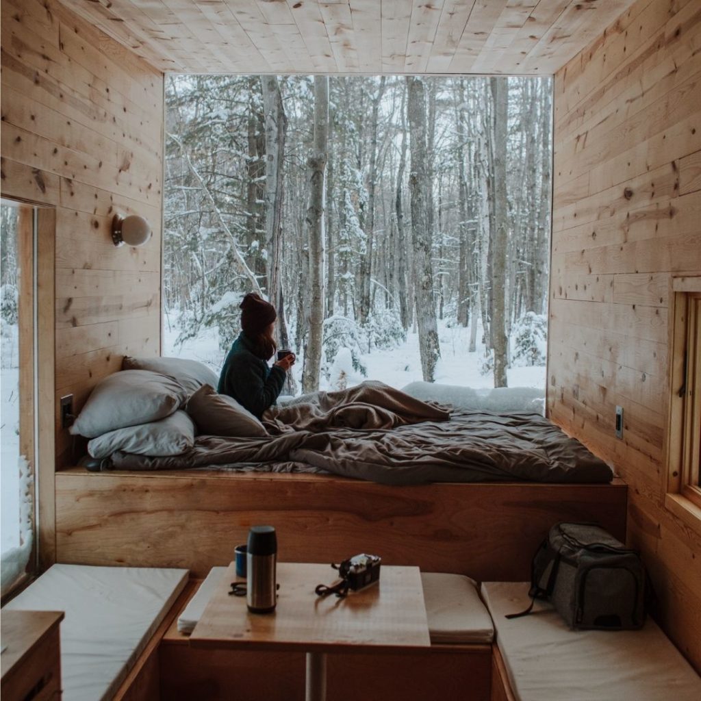A woman is curled up in bed with a cup of coffee in a wooden chalet. There's a big window in front of her, showing a snowy forest.