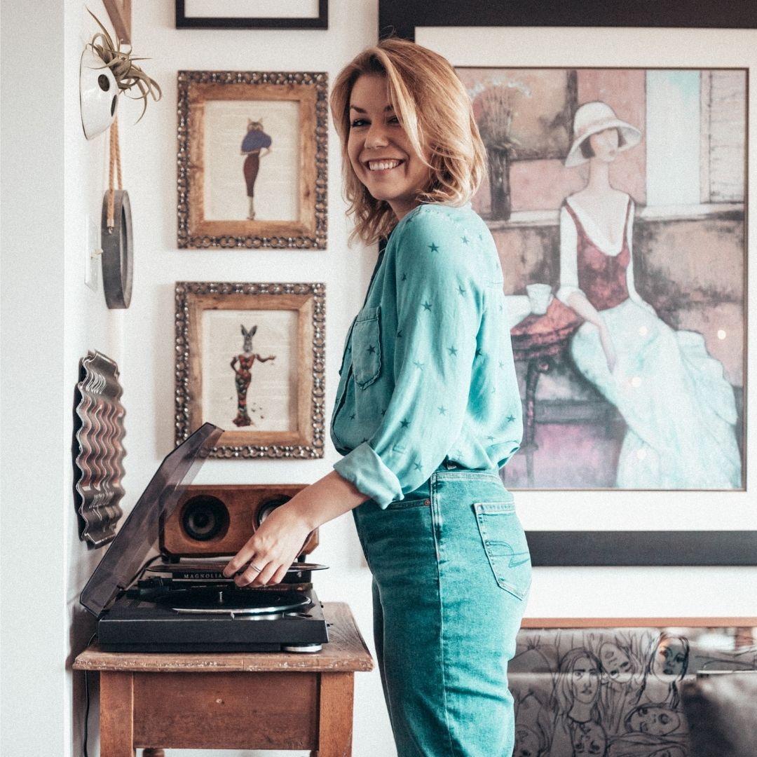 Lauren is placing a record on a record player. There is tons of art on the walls around her, and she's wearing double denim while smiling.